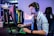 A young person is intensely focused on a computer screen while wearing a headset with a microphone. They are using a gaming keyboard and mouse, sitting in a gaming chair. The room has cool lighting, with a visible PC tower.