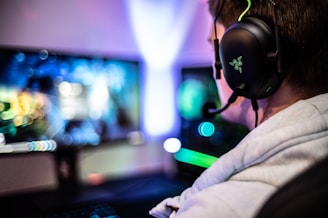 A person wearing headphones is sitting in front of a computer setup with multiple monitors displaying colorful, blurred graphics. The headphones have a distinctive logo illuminated in green. The ambient lighting and screen glow add to the immersive gaming environment.