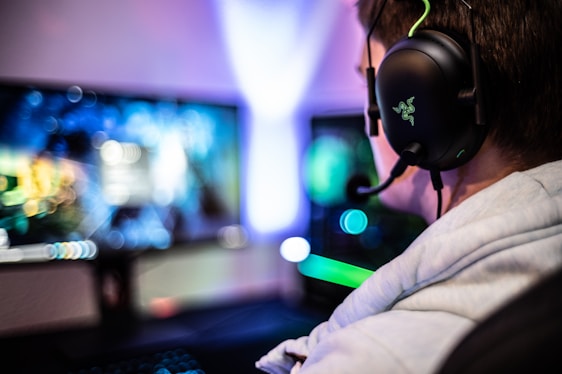 A person wearing headphones is sitting in front of a computer setup with multiple monitors displaying colorful, blurred graphics. The headphones have a distinctive logo illuminated in green. The ambient lighting and screen glow add to the immersive gaming environment.