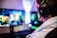 A person wearing headphones is sitting in front of a computer setup with multiple monitors displaying colorful, blurred graphics. The headphones have a distinctive logo illuminated in green. The ambient lighting and screen glow add to the immersive gaming environment.