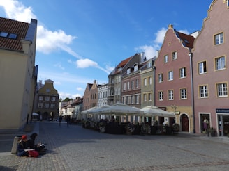 A picturesque European street lined with colorful buildings featuring ornate architectural details. Cobblestone pavement leads through the square, where outdoor cafes with white umbrellas offer seating. Two people are sitting on a ledge in the foreground, one of them engaged in writing or sketching.