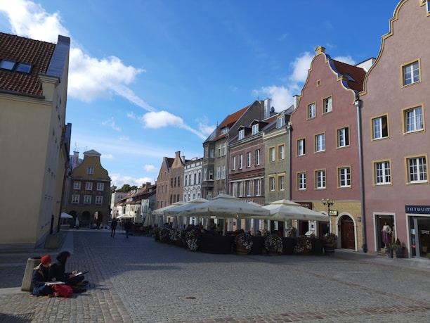 A picturesque European street lined with colorful buildings featuring ornate architectural details. Cobblestone pavement leads through the square, where outdoor cafes with white umbrellas offer seating. Two people are sitting on a ledge in the foreground, one of them engaged in writing or sketching.