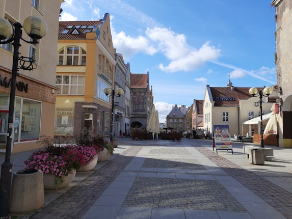 A quaint street lined with pastel-colored buildings featuring a mixture of traditional and modern architecture. Flower pots with vibrant pink and purple flowers decorate the walkway, and outdoor seating areas with umbrellas are visible in the distance. The scene is illuminated by a clear blue sky with scattered clouds.