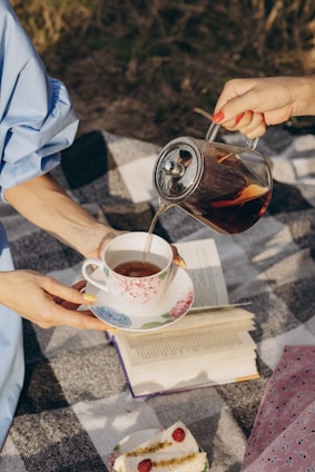 A person is pouring tea from a glass teapot into a floral-patterned teacup held by another person. A book lies open on a checkered blanket, and a plate with slices of cake garnished with raspberries is nearby.