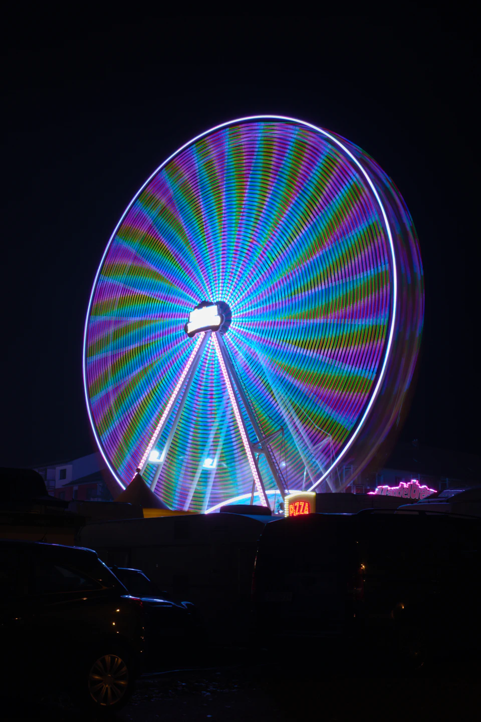 The image depicts a large, brightly lit Ferris wheel at night. The Ferris wheel is illuminated with purple and blue lights that create a vibrant contrast against the dark sky. It stands tall on a circular platform, surrounded by various vehicles parked in front of it. In the background, there are several cars and a truck, adding to the urban setting.