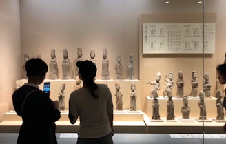 Several small, ancient sculptures are displayed on shelves within a glass case in a museum. The sculptures depict figures in various poses, some holding objects or gesturing. People in the foreground are observing and photographing the artifacts.