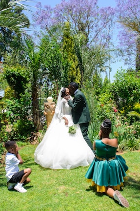 A wedding scene takes place in a lush garden with a couple kissing in the center. The bride wears a white gown and veil, while the groom is in a dark suit. Two children, one boy and one girl, are kneeling nearby. The boy covers his eyes, and the girl appears to be smiling with her hands on her cheeks. The background is filled with vibrant greenery and purple flowering trees.