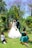 A wedding scene takes place in a lush garden with a couple kissing in the center. The bride wears a white gown and veil, while the groom is in a dark suit. Two children, one boy and one girl, are kneeling nearby. The boy covers his eyes, and the girl appears to be smiling with her hands on her cheeks. The background is filled with vibrant greenery and purple flowering trees.