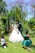 A wedding scene takes place in a lush garden with a couple kissing in the center. The bride wears a white gown and veil, while the groom is in a dark suit. Two children, one boy and one girl, are kneeling nearby. The boy covers his eyes, and the girl appears to be smiling with her hands on her cheeks. The background is filled with vibrant greenery and purple flowering trees.