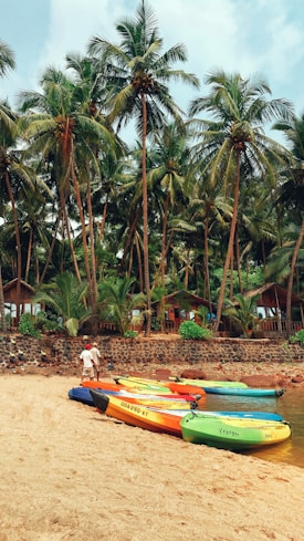 A scenic beach setting with multiple colorful kayaks resting on the sand. Tall, lush palm trees line the background, and a couple of wooden huts are partially visible among the trees. Two people stand near the kayaks, appearing to be readying them for use.
