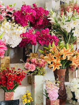 A vibrant display of various artificial flowers arranged in decorative vases, featuring bright pink orchids, white lilies, orange lilies, pink roses, red blossoms, and yellow flowers.
