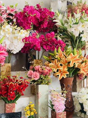 A vibrant display of various artificial flowers arranged in decorative vases, featuring bright pink orchids, white lilies, orange lilies, pink roses, red blossoms, and yellow flowers.