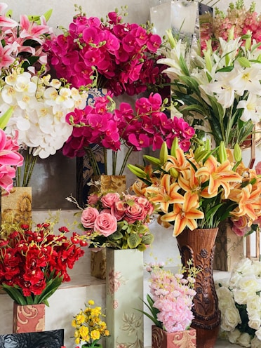 A vibrant display of various artificial flowers arranged in decorative vases, featuring bright pink orchids, white lilies, orange lilies, pink roses, red blossoms, and yellow flowers.