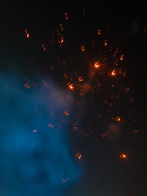 Sparks of bright orange light dance against a dark background, contrasting with a plume of blue smoke swirling upward. The scene conveys movement and energy, with the glowing particles creating a dynamic visual effect.