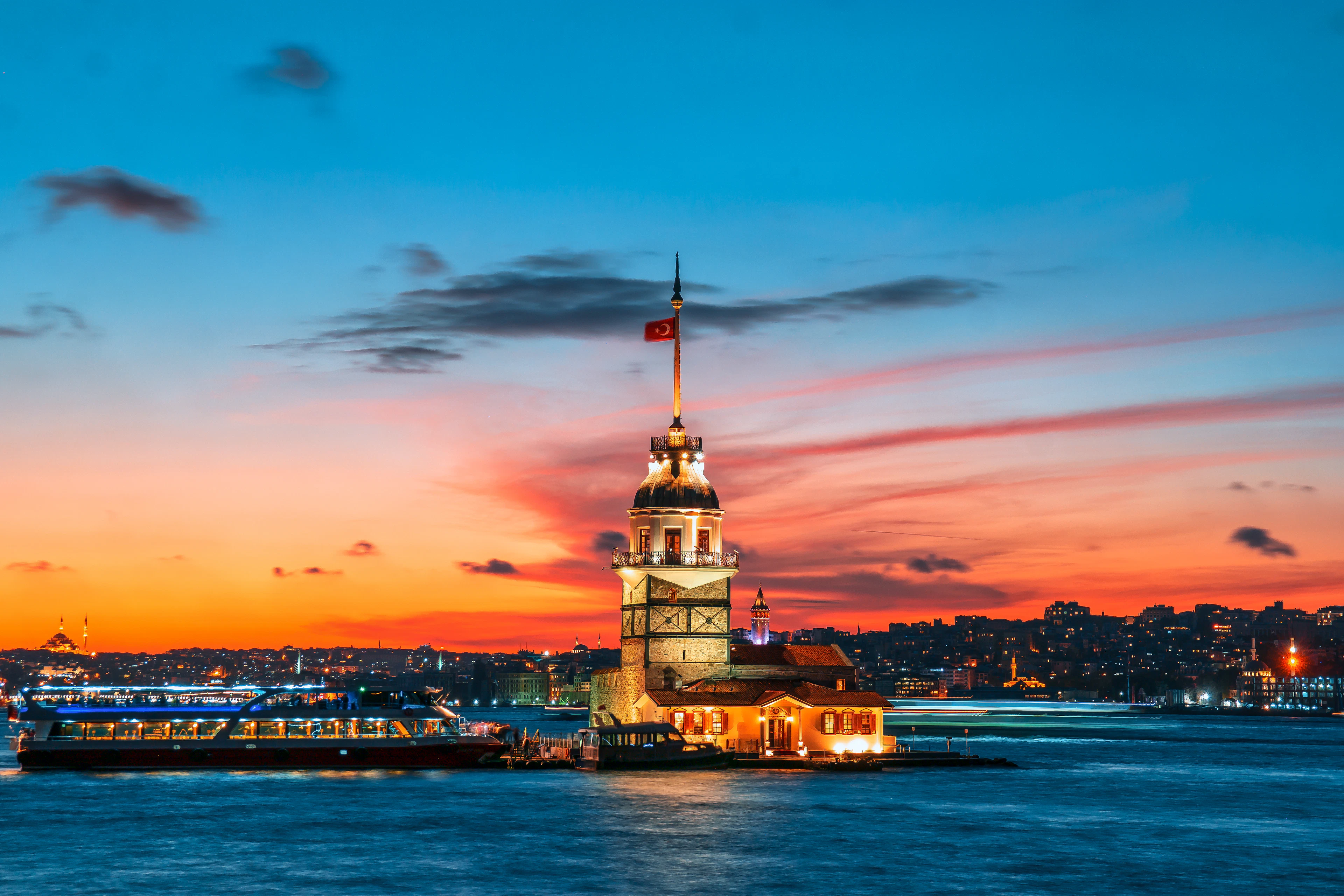Maiden's Tower lit up against a deep blue sky with a fading orange sunset band reflected on the water.