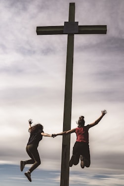 Two people are holding hands and jumping joyfully beside a large cross structure, set against a backdrop of a cloudy sky. Their silhouettes contrast with the bright sky, conveying a sense of freedom and excitement.