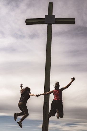 Two people are holding hands and jumping joyfully beside a large cross structure, set against a backdrop of a cloudy sky. Their silhouettes contrast with the bright sky, conveying a sense of freedom and excitement.