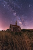 A dark, starry night sky featuring the Milky Way stretches over an old, rustic church with a spire. The church is surrounded by tall, dry grass, and the scene is illuminated by a soft glow. Shooting stars are visible in the sky, adding to the celestial atmosphere.