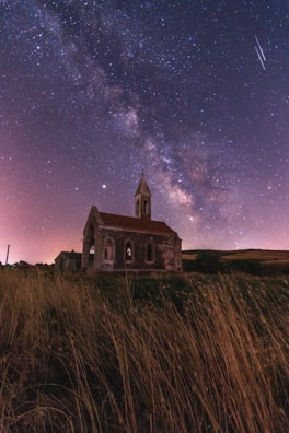 A dark, starry night sky featuring the Milky Way stretches over an old, rustic church with a spire. The church is surrounded by tall, dry grass, and the scene is illuminated by a soft glow. Shooting stars are visible in the sky, adding to the celestial atmosphere.