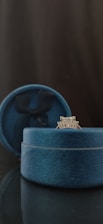 A silver ring with a diamond setting is displayed in an open blue velvet ring box. The box is set against a dark background, with an elegant and sophisticated appearance.