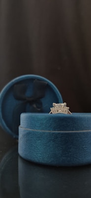A silver ring with a diamond setting is displayed in an open blue velvet ring box. The box is set against a dark background, with an elegant and sophisticated appearance.