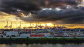 An industrial port with cranes unloading metal containers at sunset.