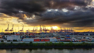A dimly lit industrial port at dusk with silhouettes of cranes and containers under a charcoal sky.