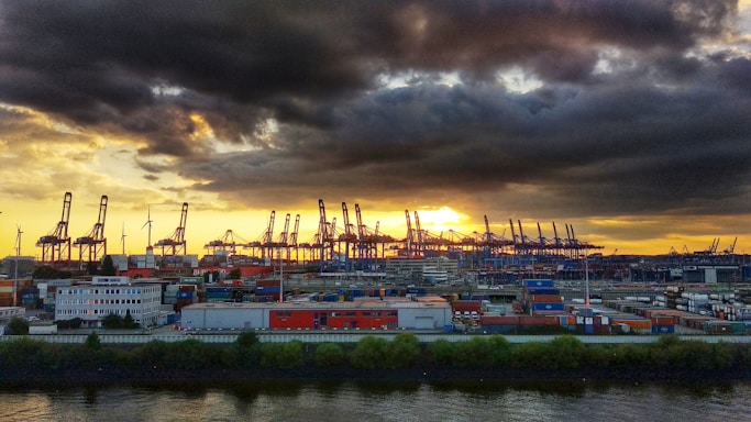 A vibrant port scene at sunset with shipping containers and cranes silhouetted against the sky.