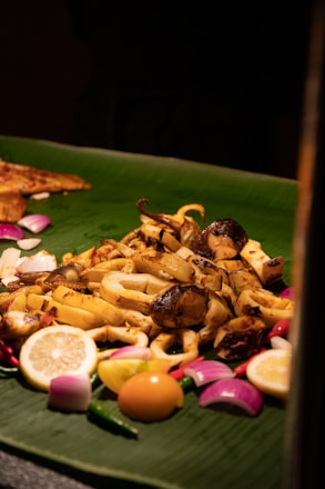 A platter of assorted grilled seafood placed on a large green banana leaf. It is garnished with lemon slices, red onions, green chili peppers, and a whole egg yolk. The seafood appears to be seasoned and cooked with a slightly charred look.
