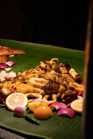 A platter of assorted grilled seafood placed on a large green banana leaf. It is garnished with lemon slices, red onions, green chili peppers, and a whole egg yolk. The seafood appears to be seasoned and cooked with a slightly charred look.