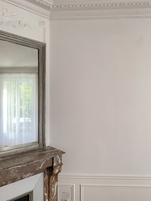 An elegant interior space featuring intricate wall moulding and a large mirror above a stone fireplace. The walls are light-colored with detailed decorative trim along the ceiling. A curtain filters natural light from a window partially visible in the mirror.