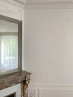 An elegant interior space featuring intricate wall moulding and a large mirror above a stone fireplace. The walls are light-colored with detailed decorative trim along the ceiling. A curtain filters natural light from a window partially visible in the mirror.