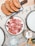 Slices of cured meat arranged on a white plate, next to a cutting board with several pieces of rustic bread topped with tomato. A cocktail with a straw and sprigs of rosemary is visible, as well as a glass with water. The table is covered with a paper mat featuring printed words and phrases.
