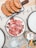 Slices of cured meat arranged on a white plate, next to a cutting board with several pieces of rustic bread topped with tomato. A cocktail with a straw and sprigs of rosemary is visible, as well as a glass with water. The table is covered with a paper mat featuring printed words and phrases.