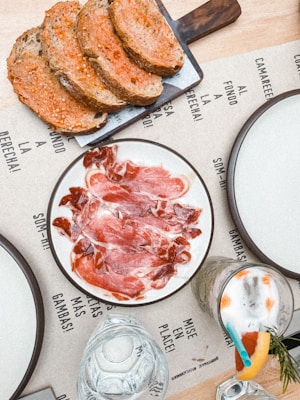 Slices of cured meat arranged on a white plate, next to a cutting board with several pieces of rustic bread topped with tomato. A cocktail with a straw and sprigs of rosemary is visible, as well as a glass with water. The table is covered with a paper mat featuring printed words and phrases.