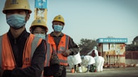 A group of construction workers wearing orange safety vests, yellow helmets, and blue masks stand in a line. They appear to be on a construction site with a crane and other workers in white protective suits in the background. The setting suggests a mixture of construction and possibly some form of safety or health protocol.