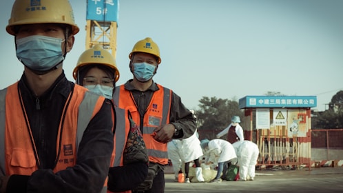 A group of construction workers wearing orange safety vests, yellow helmets, and blue masks stand in a line. They appear to be on a construction site with a crane and other workers in white protective suits in the background. The setting suggests a mixture of construction and possibly some form of safety or health protocol.