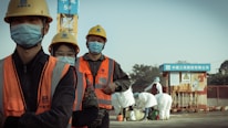 A group of construction workers wearing orange safety vests, yellow helmets, and blue masks stand in a line. They appear to be on a construction site with a crane and other workers in white protective suits in the background. The setting suggests a mixture of construction and possibly some form of safety or health protocol.