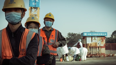 A group of construction workers wearing orange safety vests, yellow helmets, and blue masks stand in a line. They appear to be on a construction site with a crane and other workers in white protective suits in the background. The setting suggests a mixture of construction and possibly some form of safety or health protocol.