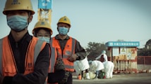 A group of construction workers wearing orange safety vests, yellow helmets, and blue masks stand in a line. They appear to be on a construction site with a crane and other workers in white protective suits in the background. The setting suggests a mixture of construction and possibly some form of safety or health protocol.