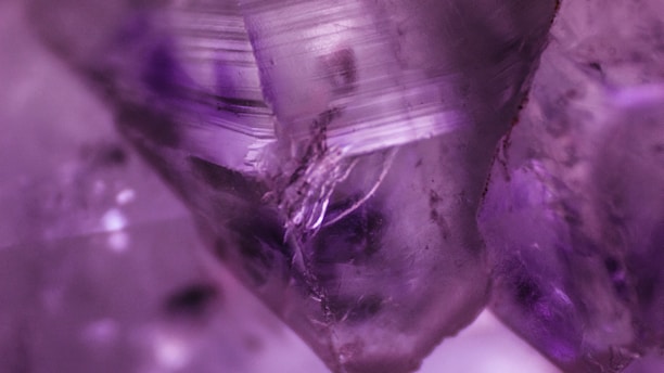 A close-up view of a purple crystalline structure with intricate patterns and textures. The crystal appears translucent with areas of deeper violet hues, showcasing natural inclusions and striations on its surface.