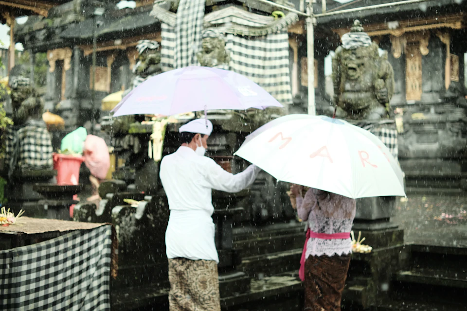 Two people walking under umbrellas through a Bali street during a December wet-season rain shower