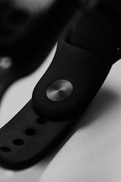 Close-up of a black wristband with a metallic button. The focus is on the circular button, highlighting its texture and the smooth, matte surface of the band. Light reflects subtly off the metal and the black material, creating a sleek and modern appearance.