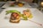 A gourmet dish is presented on a white square plate, featuring a beautifully cooked piece of fish topped with a tomato-based garnish and asparagus spears. Beside the fish, there are two grilled shrimp sitting on a bed of corn relish with a drizzle of red sauce adding a pop of color. Another plate with different food items is visible in the background.