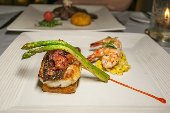 A gourmet dish is presented on a white square plate, featuring a beautifully cooked piece of fish topped with a tomato-based garnish and asparagus spears. Beside the fish, there are two grilled shrimp sitting on a bed of corn relish with a drizzle of red sauce adding a pop of color. Another plate with different food items is visible in the background.