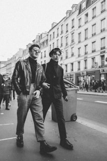 Two men in fashionable attire are walking on a city street, surrounded by tall buildings with classic architecture. The image is in black and white, adding a vintage feel. One man is wearing a leather jacket and checkered pants, while the other is dressed in a dark coat and trousers. Other pedestrians can be seen in the background, contributing to the bustling city atmosphere.