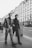 Two men in fashionable attire are walking on a city street, surrounded by tall buildings with classic architecture. The image is in black and white, adding a vintage feel. One man is wearing a leather jacket and checkered pants, while the other is dressed in a dark coat and trousers. Other pedestrians can be seen in the background, contributing to the bustling city atmosphere.