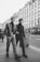 Two men in fashionable attire are walking on a city street, surrounded by tall buildings with classic architecture. The image is in black and white, adding a vintage feel. One man is wearing a leather jacket and checkered pants, while the other is dressed in a dark coat and trousers. Other pedestrians can be seen in the background, contributing to the bustling city atmosphere.