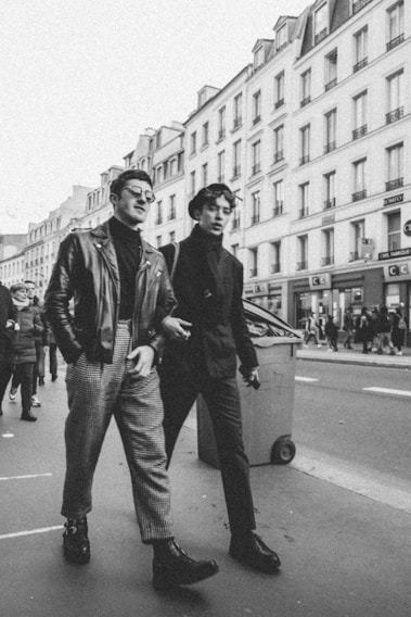 Two men in fashionable attire are walking on a city street, surrounded by tall buildings with classic architecture. The image is in black and white, adding a vintage feel. One man is wearing a leather jacket and checkered pants, while the other is dressed in a dark coat and trousers. Other pedestrians can be seen in the background, contributing to the bustling city atmosphere.