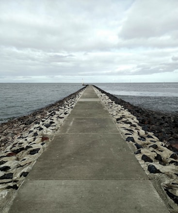 A long, narrow pathway stretches into the distance, bordered by rocks on both sides, leading into the sea under a cloudy sky. The pathway is made of concrete, and a small structure is visible at the end of it.